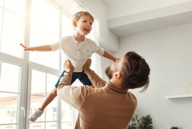 Neşeli genç baba heyecanlı küçük çocuğu tutuyor birlikte vakit geçiriyor ve evde ışık odasında eğleniyor.