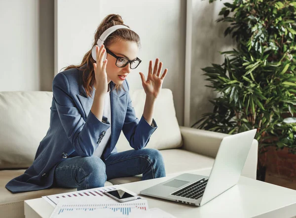 Serious young female in casual outfit and glasses having video conversation with colleagues and sharing business ideas while sitting on couch in modern workplac