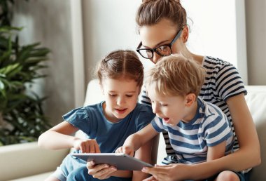 Küçük bir oğlu ve kızı olan neşeli genç bir anne evde birlikte vakit geçirirken kanepede oturuyor ve tablet üzerinde video oyunu oynuyor.