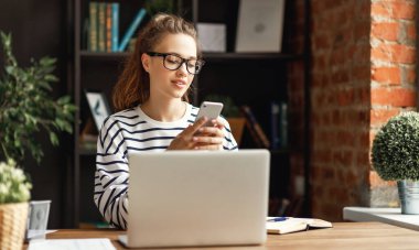 Genç bir kadın dizüstü bilgisayarda çalışıyor ve akıllı telefonunda bilgi arıyor. A masasında oturuyor ve evdeki uzak proje üzerinde çalışıyor.