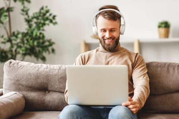 快乐的大胡子男人 带着手提电脑 头戴无线耳机 穿着休闲装 一个人坐在柔软的沙发上 愉快地听着轻盈宽敞的客厅里最喜欢的音乐 — 图库照片