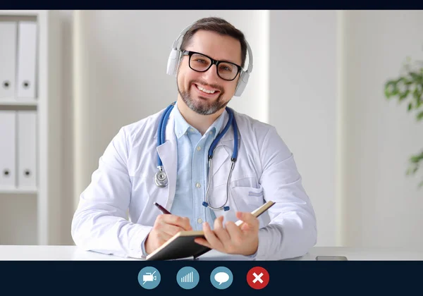 Happy medical practitioner in headphones smiling for camera and writing in notebook during video cha