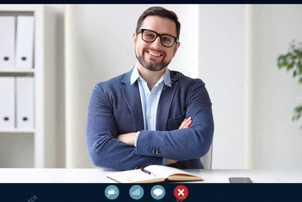 Confident businessman with crossed arms smiling and looking at camera while sitting in office and attending online meetin