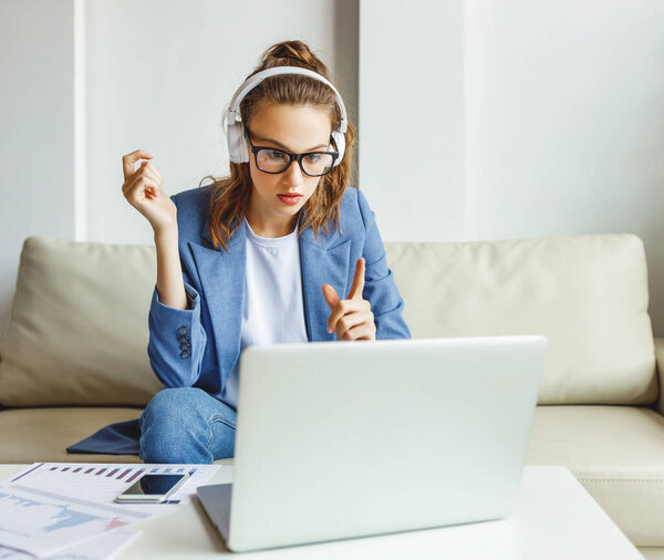 Serious young female in casual outfit and glasses having video conversation with colleagues and sharing business ideas while sitting on couch in modern workplac