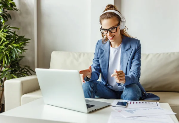 Cheerful young female in casual outfit and glasses having video conversation with colleagues and sharing business ideas while sitting on couch in modern workplac