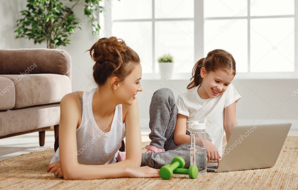 Happy young female and little daughter in sportswear sitting on floor near laptop and watching video tutorial while preparing for workout together at hom