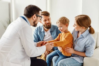 Modern klinikteki rahat bir muayenehanede muayene sırasında mutlu çocuk ve ebeveynlerle konuşan pozitif erkek çocuk doktoru. 