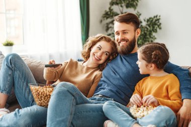 İyimser bir aile, gündelik kıyafetleriyle, ışık odasında kanepede oturuyor ve TV izlerken gülüyor ve güneşli bir günde lezzetli patlamış mısır yiyor.
