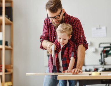 Heyecanlı küçük çocuk ve benzer gömlekler giyen genç baba modern el sanatları Studi 'sinde ahşapla çalışırken bir tahtayı testereyle kesiyorlar.