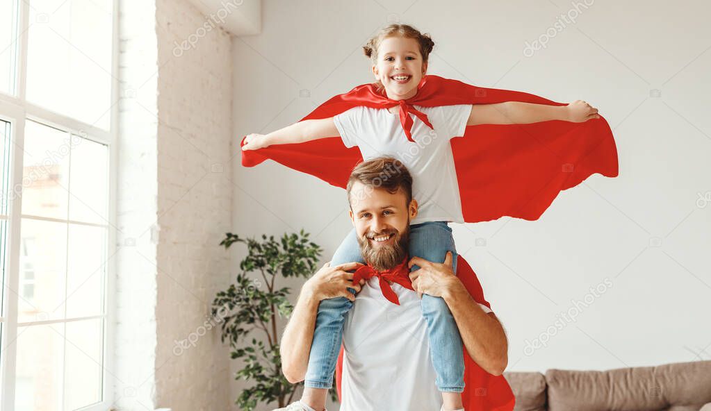 Smiling bearded young man holding cute girl on shoulders while playing superheroes together dressed in red superhero cloak in light roo