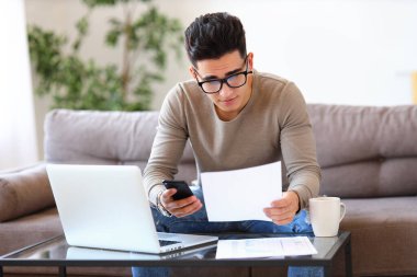 Gözlüklü etnik adam, kanepede otururken ve evdeki evrak işlerini yaparken belgelerden veri toplamak için akıllı telefon kullanıyor.