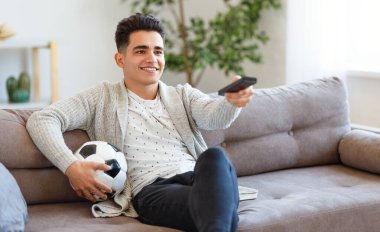 Gülen ve uzaktan kumanda kullanan topları olan mutlu etnik genç adam hafta sonları evde oturup futbol maçı izliyor.