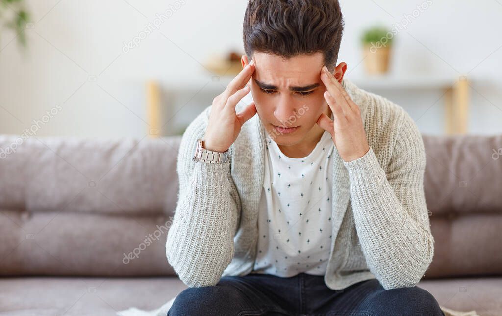 Stressed young ethnic guy in casual clothes sitting on sofa and rubbing temples while suffering from migraine at hom