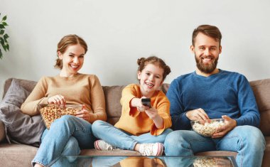 İyimser bir aile, gündelik kıyafetleriyle, ışık odasında kanepede oturuyor ve TV izlerken gülüyor ve güneşli bir günde lezzetli patlamış mısır yiyor.