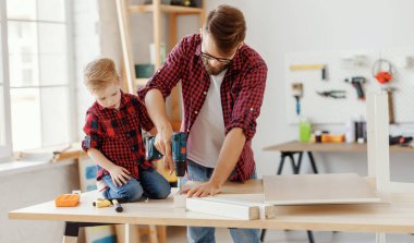 Heyecanlı küçük çocuk ve genç baba benzer gömlekler içinde modern el işi Studi 'de odunla çalışırken matkabı bir arada tutuyorlar.