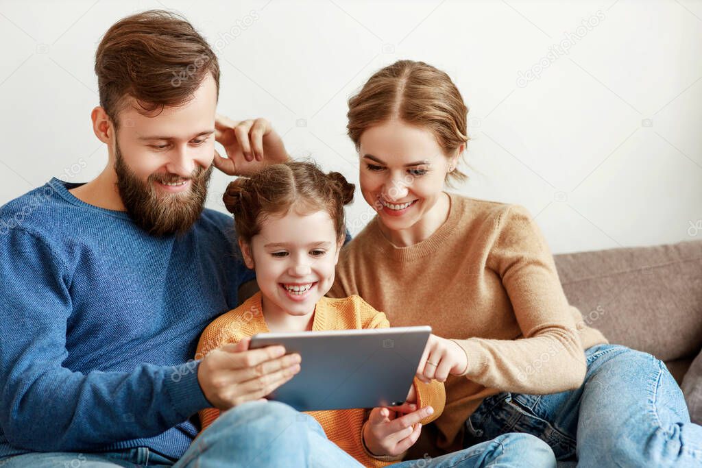 Optimistic young parents with laughing daughter in casual outfit sitting on couch browsing tablet while spending time together during sunny da