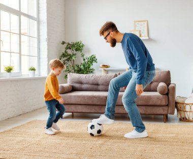 Uzun boylu neşeli çocuk evde futbol oynarken sakallı babasının yanında gülüyor.