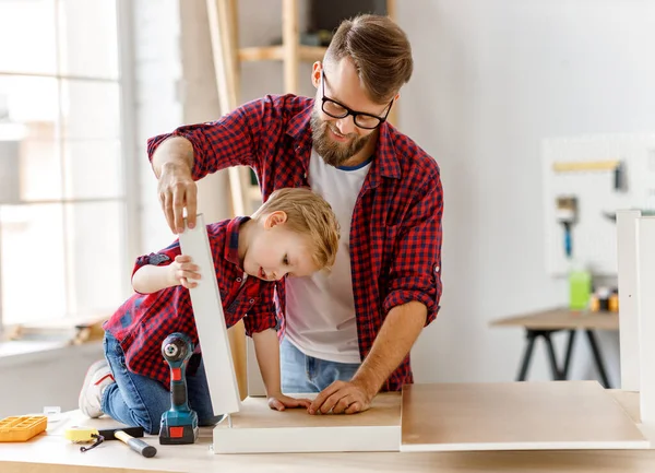 Delighted little boy in casual clothes laughing and helping father to   assembling furniture on table in home workshop togethe