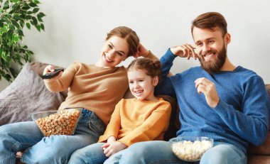 İyimser bir aile, gündelik kıyafetleriyle, ışık odasında kanepede oturuyor ve TV izlerken gülüyor ve güneşli bir günde lezzetli patlamış mısır yiyor.
