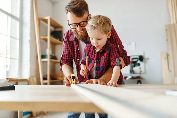 Delighted little boy in casual clothes laughing and helping father to   assembling furniture  and measure the board on table in home workshop togethe
