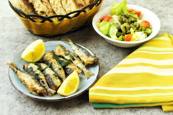 Limon ve maydanozla süslenmiş tabakta kızarmış sardalya, salata kasesi ve Arap ekmeği.