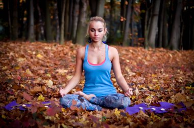 Mavi tişörtlü genç sarışın kadın, sonbahar ormanında kapalı gözlerle meditasyon yapan yarı lotus pozisyonunda yerde oturarak yoga yapıyor. Yerde yatan sarı ve kırmızı yapraklar.
