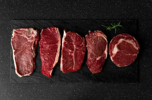 Selection of raw beef meat food steaks against black stone background.