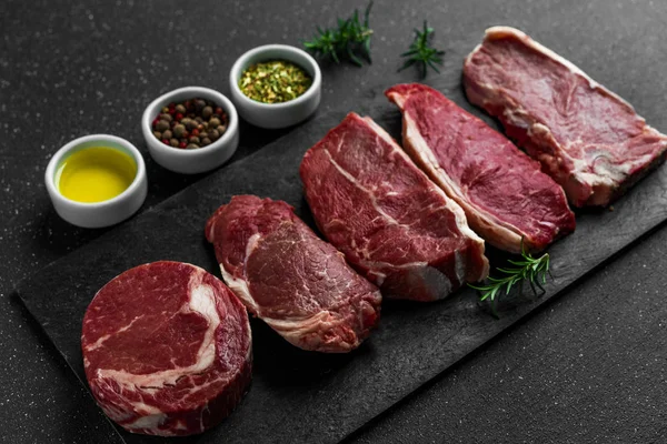 Selezione di carne di manzo cruda bistecche alimentari sullo sfondo di pietra nera . — Foto Stock