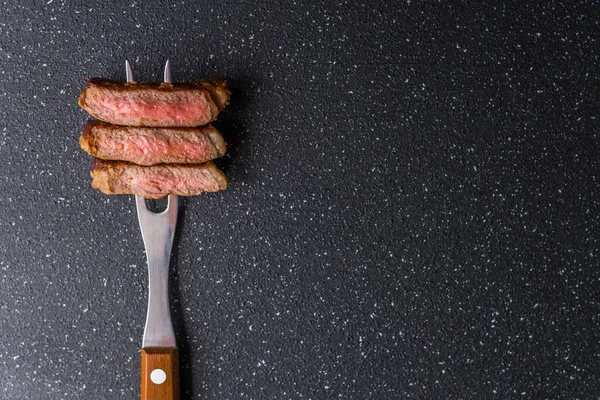 New York strip lendenen rundvlees biefstuk op een vlees vork tegen zwarte steen achtergrond. — Stockfoto