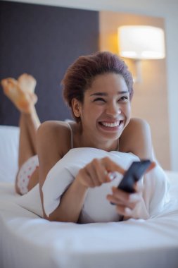 Young woman watching TV in the room. Young beautiful woman in bed. Young black woman seat in bed. clipart