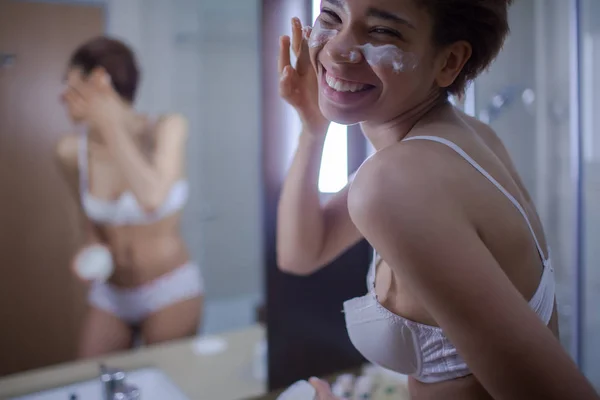Krásná mladá Afričanka šíření krém na obličej a usmívá se stojící před zrcadlem v koupelně — Stock fotografie