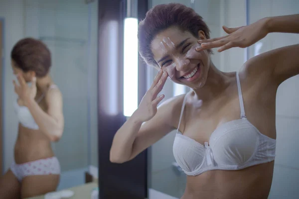 Beautiful young African woman spreading cream on her face and smiling standing against a mirror in bathroom