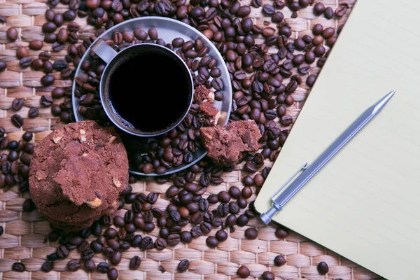 Xícara de café e grãos de café em mesa de madeira com biscoitos e bloco de notas — Fotografia de Stock