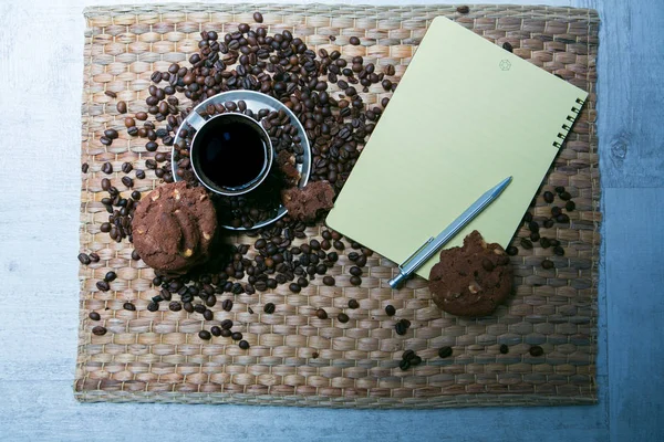 Taza de café y granos de café en la mesa de madera con galletas y bloc de notas — Foto de Stock