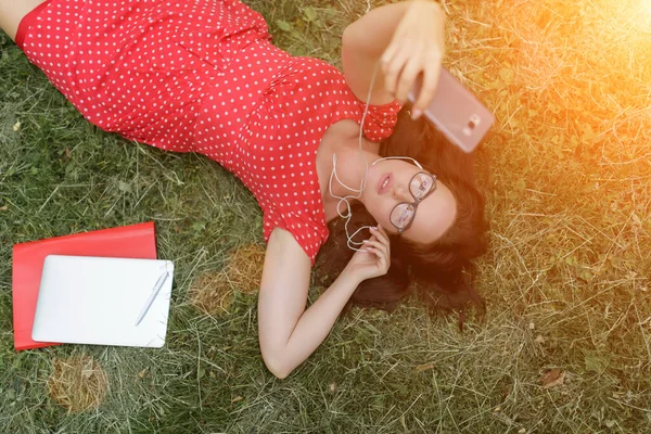 Mídia, entretenimento musical. Transmissão de música. Mulher relaxante na grama verde. Morena ouvir música, tomando selfie — Fotografia de Stock