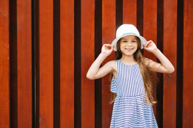 Çocuklar, yüz ifadeleri, duygular, moda kavramı. Beyaz şapka ve kameraya poz onun yeni modern giyimleri gösteren çizgili elbise giyen gülümseyen mutlu kız çekici görünümü ile