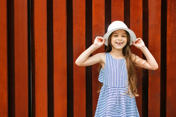 Lovely girl in summer white hat and striped dress, having broad smile, keeping her hands on hat posing against fence background with copy space for your text. Summer girl with good mood resting