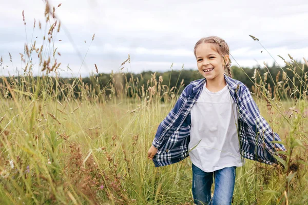 Criança pequena bonita com pigtail, vestindo camisa xadrez e jeans, sorrindo agradavelmente enquanto corre na grama verde dos prados. Criança feminina feliz descansando ao ar livre, se divertindo e bom humor — Fotografia de Stock