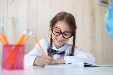 Hard-working little schoolgirl with two pigtails, wearing big glasses and school uniform, doing her homework while sitting at table, writing in copy book with happy expression. Children, education clipart