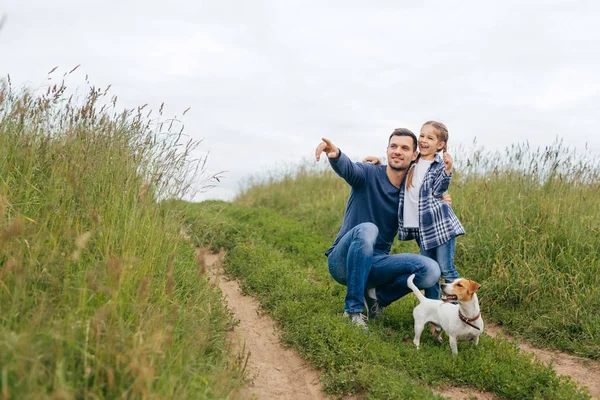 Menina bonita, seu pai e seu cão andando juntos através do campo verde, admirando belas paisagens e se divertindo juntos. Pai mostrando sua filha floresta apontando com o dedo indicador — Fotografia de Stock