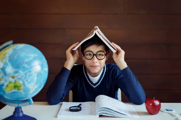 Busy schoolboy, doing his home assigment, keeping book on head, looking puzzled while not knowing how to do exercise. Boy sitting at his work place, surrounded with books and school equipment