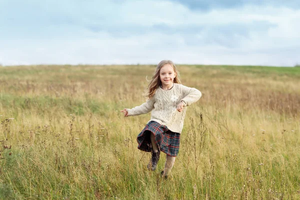 Niña hermosa vistiendo suéter caliente y falda, corriendo en el prado verde, admirando el aire fresco, el cielo encantador y la naturaleza. Una niña bonita posando en el campo. Niños, relajación, concepto de naturaleza — Foto de Stock