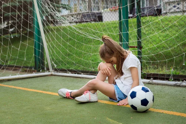 Little active girl wearing T-shirt, jean shorts and running shoes, hanging her head while being upset to miss goal in gates. Unhappy female kid goalkeeper playing football, crying because of defeat