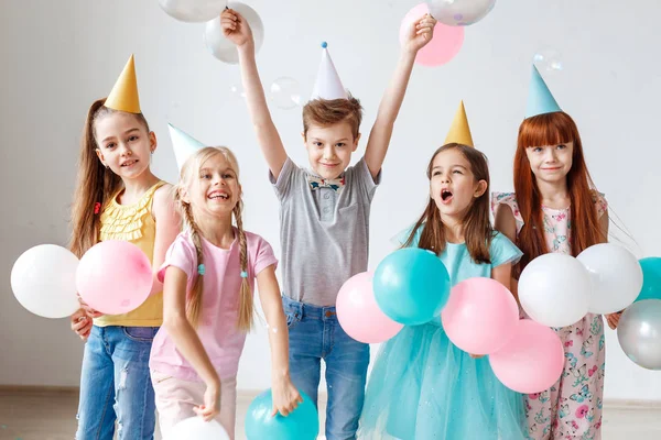 Grupo de niños pequeños tienen fiesta de cumpleaños, usan sombreros festivos, sostienen globos, tienen alegría juntos, disfrutan jugando juegos. Pequeña chica adorable celebra su cumpleaños, invita a amigos, tienen miradas felices —  Fotos de Stock