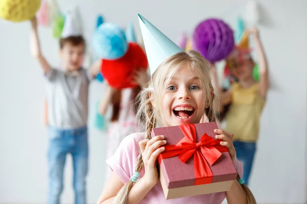 Divertido sonriente hermoso niño pequeño lleva sombrero de fiesta, abraza la caja envuelta grande, contento de recibir regalo de familiares, celebra el cumpleaños con amigos, los niños pequeños se divierten en el fondo —  Fotos de Stock