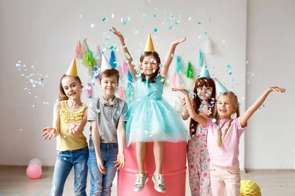 Los niños adorables se divierten juntos, lanzan confeti colorido, usan sombreros de cono, se divierten en la fiesta de cumpleaños, juegan juntos en diferentes juegos, pasan tiempo en la habitación decorada. Concepto de niños y eventos —  Fotos de Stock