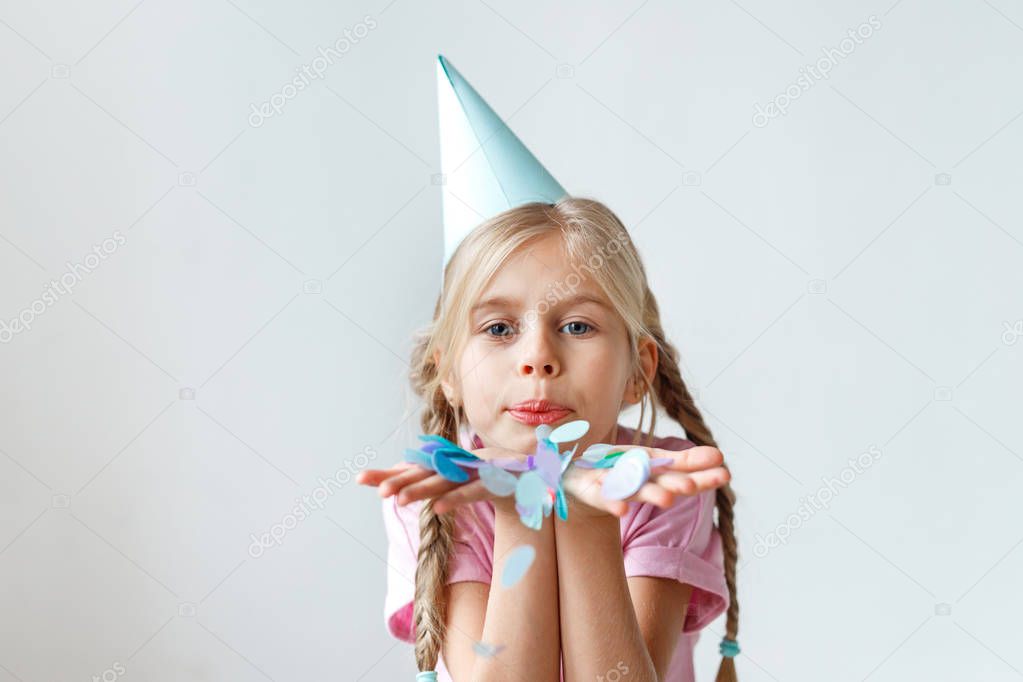 Indoor shot of pretty small kid with long pigtails, wears cone hat on head, blows colourful sheet of paper, looks into camera, celebrates her anniversary, isolated over white studio background