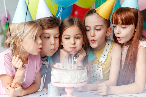 Festliche Kinder in Partymützen, Kerzen auf leckeren Kuchen pusten, Wünsche machen, Geburtstag feiern, gemeinsam feiern, bunte Luftballons in die Höhe halten. glückliches kleines Mädchen verbringt festliches Ereignis mit besten Freunden — Stockfoto