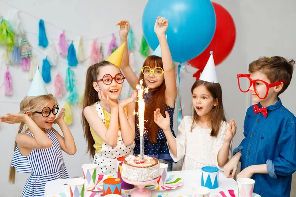 Indoor shot of happy joyful children look at big sparkle on cake, celebrar el cumpleaños, llevar gafas grandes incómodas, gorras de fiesta, estar cerca de la mesa con dulces en la habitación decorada, tienen expresiones excitadas —  Fotos de Stock