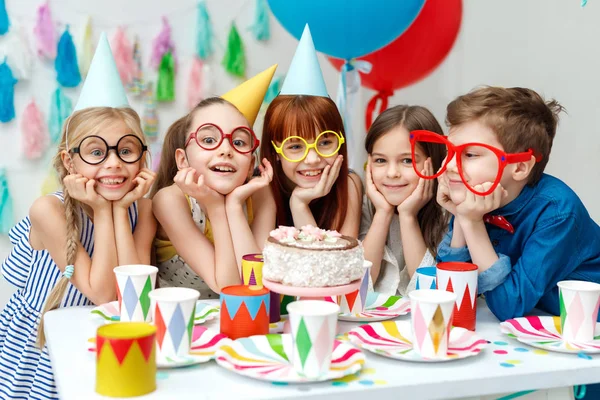 Retrato de grupo divertido de niños usar gorras de fiesta, grandes espectáculos, mirar con gran apetito en pastel de cumpleaños, quiere probarlo, tener ocasión especial, pasar tiempo felizmente. Celebración, concepto de vacaciones —  Fotos de Stock